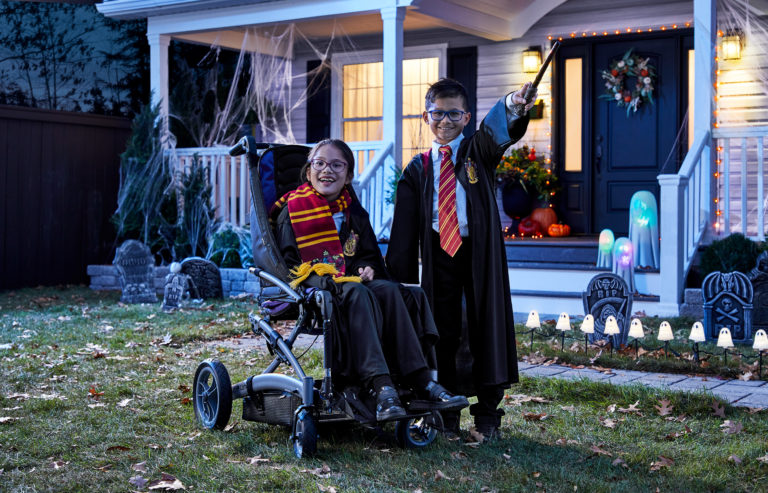 Two children dressed up in Harry Potter character costumes. The child on the left is using a wheelchair. They are posed in front of a house that's decorated with spiderwebs and tombstones.